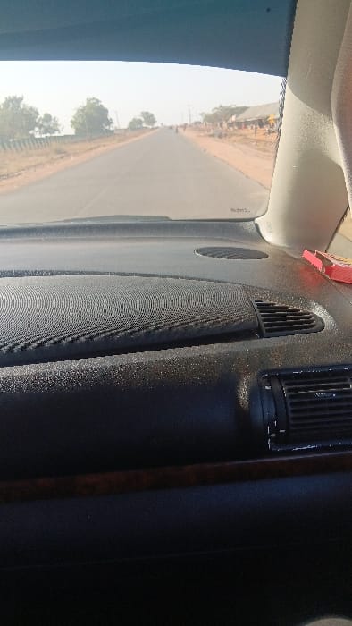 The image provides a view from inside a vehicle, looking out through the front passenger side window and across the dashboard. The scene is oriented vertically, suggesting the photo was taken with a phone in portrait mode and rotated.
The dashboard, which occupies a significant portion of the foreground, is dark-colored (likely black or dark grey) with a slightly textured or speckled appearance, possibly from dust or the material itself. It features several components:
* Two air vents with vertical slats are visible on the dashboard, one near the center and another closer to the passenger door.
* A large, rectangular speaker grille with a fine mesh pattern is centrally located on the dashboard.
* A thin, dark reddish-brown trim piece, possibly wood-grain, runs vertically along the right side of the dashboard, bordering the darker material.
* Near the windshield, above the central dashboard, a small, reddish object, possibly an air freshener or tag, is attached or resting. Faint, indistinct text in a lighter color can be seen on it, but it's not legible.
Outside the vehicle, a paved, two-lane road is visible, extending into the distance under a bright, somewhat hazy daytime sky. The road is flanked by dirt shoulders, and further beyond, there are scattered green trees and vegetation. The overall impression of the external environment is rural or semi-rural.
A noticeable reflection is present on the windshield or a side window in the upper left portion of the image. This reflection superimposes an image of the road ahead, showing distant figures or structures along the roadside, and possibly a small yellow object or vehicle in the far distance.
The vehicle appears to be in motion, driving along the road. The time of day is clearly daytime, characterized by bright, diffused light. No specific location or additional activities are discernable. No clear, legible text is visible in the image.