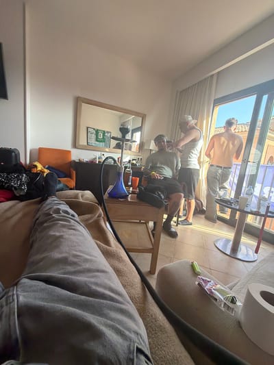 The image shows an indoor scene, likely a hotel room in Punta Umbría, Spain. Three men are present near a glass door or window leading to a balcony, where part of a building exterior can be seen. One man is shirtless, another is in a tank top and hat, and the third man is seated while the second man appears to be grooming him. A table is positioned nearby. In the foreground, a person, presumably the photographer, is sitting on a couch, with the lower part of their leg visible. A hookah is set up on a small table adjacent to the couch. Personal items, including a bag and clothes, are scattered near an orange chair. A mirror hangs on the wall, reflecting items on a shelf. A roll of toilet paper is also visible.