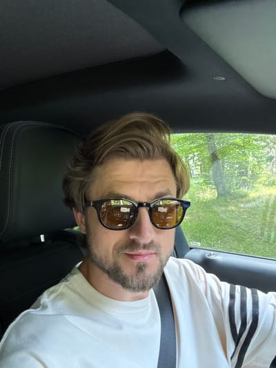 The image shows a close-up, portrait-style shot of a man wearing sunglasses and a white t-shirt with black stripes on the sleeves. He is inside a vehicle, with the interior upholstery and a window with greenery visible in the background. The man has light brown hair and a beard. He appears to be looking towards the camera, with his head tilted. The scene suggests a casual, possibly daytime setting, given the natural light and the greenery outside. The image is oriented sideways. There is no visible text in the image. The location context provided is Krubki-Górki, Poland.