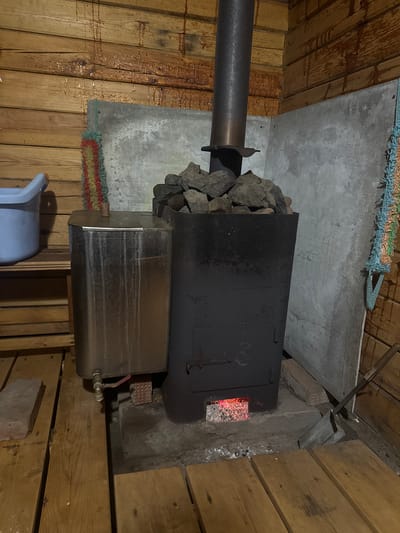 The image depicts a sauna stove in a wooden structure, likely a sauna room. The stove is black and contains rocks on top, presumably for heat retention. The firebox is partially visible, showing glowing embers inside. A metal pipe extends from the stove, likely for ventilation. Below the stove is a water heater, also made of metal. A plastic bucket sits on the floor near the heater. The wooden walls of the sauna are visible on either side, and a concrete or stone wall is behind the stove. Based on the prompt, the location is likely in the city of Mayak, Russia.