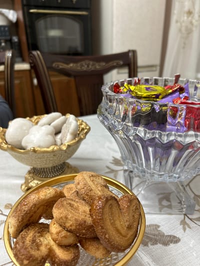 In the city of Nizhnee Kazanishche, Russia, a table is set with three bowls of sweets. In the foreground, a golden-rimmed bowl holds palmiers, a type of puff pastry cookie dusted with sugar. To the left, a decorative, ornate bowl displays white, soft sweets, possibly cookies or candies. A large, crystal glass bowl sits to the right, filled with individually wrapped chocolates in various colors, including purple, yellow, and red. The table is covered with a white tablecloth featuring a subtle floral pattern. In the background, a dark wooden dining chair is partially visible, and behind it, a black oven is mounted above kitchen cabinetry. The lighting suggests an indoor setting, likely a home dining area.