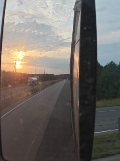 The image depicts a view from a vehicle's side mirror, reflecting a rural road scene at sunset. The setting sun casts a warm glow on the sky, with scattered clouds. A tanker truck is visible on the road, driving away from the viewer. The road is bordered by trees and fields. Power lines are strung across the sky. The mirror frame itself is visible in the foreground, obscuring a portion of the reflection. The location is identified as Chaadaevka, Russia.