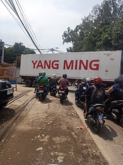 A line of motorbikes waits behind a large truck with "YANG MING" in bold red lettering. The truck's container bears the code "YNIU 5530/2 7 45P.1". The road is unpaved and uneven. Buildings and trees line the sides of the road, and electrical wires stretch overhead. A black car is parked near a sign that reads "TERSEDIA DI S" with additional text. A license plate with "D 1727 RH" is visible on the car.