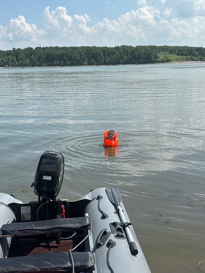 A young child wearing an orange life vest floats in a body of water. The child is facing forward and appears to be smiling. The water is calm with ripples. In the foreground, a portion of a gray inflatable boat is visible, featuring black seats and a black outboard motor. The background shows a dense line of green trees on a distant shore under a blue sky with white clouds. The location is the Neman River near the city of Pravda, Russia.