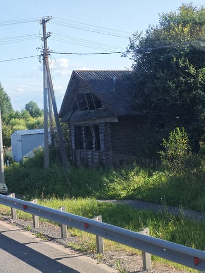 The image shows a weathered, abandoned log cabin with a corrugated metal roof, partially obscured by overgrown vegetation and a large tree. In the foreground, a section of a metal guardrail runs parallel to a paved surface, likely a road. To the left of the cabin, a light blue metal shed stands amidst the greenery. A utility pole with multiple wires is visible to the left of the cabin. The sky above is clear blue with a few wispy clouds. The scene appears to be in a rural or semi-rural setting, possibly on the outskirts of Bol'shaya Kiselenka, Russia. No people are present in the image.