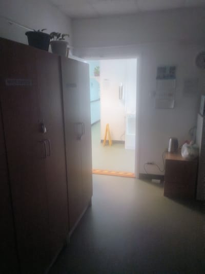 The image shows a dimly lit interior hallway. On the left, there are two tall wooden cabinets with handles and locks. The top of the left cabinet has two potted plants. Above the plants, there is a sign that is difficult to read but appears to have Cyrillic text. In the center of the image, a doorway leads to another area with bright white walls and a white appliance. A yellow A-frame sign stands in this area. On the right side of the hallway, a small wooden cabinet sits against the wall. On top of this cabinet is a silver electric kettle and a white plastic bag. A black power cord extends from the kettle to an outlet near the floor. A calendar is attached to the wall above the cabinet. The floor is a flat, neutral color. The overall scene appears to be in an institutional setting, possibly a medical facility or an office.