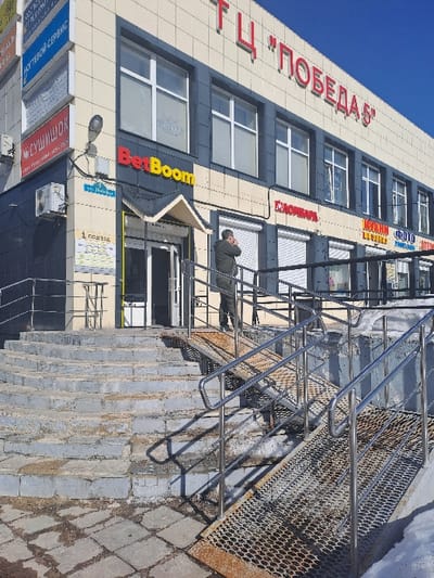 The image displays the exterior of a commercial building, specifically "Shopping Center 'Pobeda 5'" located at Lenin Street, 1A, in Yegoryevsk, Russia. The two-story building features a facade of beige and dark grey panels under a clear blue sky. Large red letters spelling "ТЦ 'ПОБЕДА 5'" are affixed to the top floor. Below, a prominent "BetBoom" sign, in red and yellow, marks a central entrance. Additional business signs for "Nail Service" and "Sushishok" are visible on the building's left corner.

Access to the main entrance is facilitated by a wide, worn set of concrete stairs and, to their right, two parallel, rusted perforated metal ramps with shiny chrome handrails. Patches of snow and ice are present on the ramps and along the base of the structure, indicating cold conditions. One individual, wearing a dark-colored hooded jacket, stands on the paved landing at the top of the stairs and ramps. This person is positioned facing to the right, with their left hand raised to their ear, consistent with being engaged in a phone call. Strong sunlight casts distinct shadows from the railings and building across the entry area.