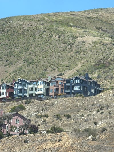 This media file depicts a scenic view of houses built on a steep, grassy hillside. The houses are clustered together, appearing to be multi-story townhouses or apartments, with varying architectural styles and colors, including dark blue, green, white, and red. They are arranged in a linear fashion along the slope of the hill. The hillside itself is covered in dry, sparse vegetation, indicating a potentially arid or Mediterranean climate. In the background, a clear blue sky suggests fair weather. The perspective is from a slightly elevated viewpoint, looking down on the houses and across the hillside. The time of day appears to be daytime, with ample sunlight casting shadows. The scene evokes a sense of residential living integrated into a dramatic natural landscape. There is no visible text in the image.
