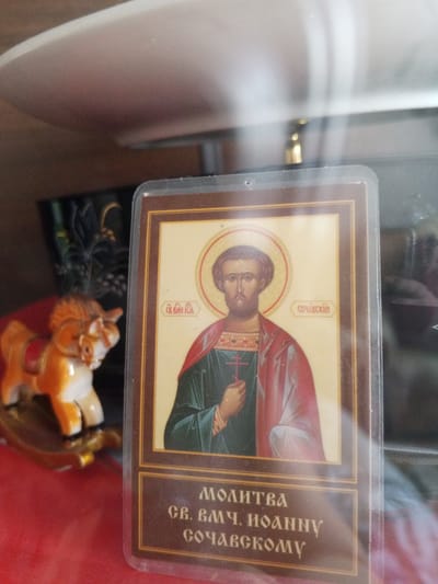 The image shows a religious icon depicting a male saint with a halo, wearing traditional robes and holding a cross. The icon is enclosed in a protective plastic sleeve. Below the image of the saint, there is text in Cyrillic script, which appears to be a prayer or invocation. To the left of the icon, a small, stylized figurine of a bull or ox, possibly a toy or decorative item, is visible. The background is blurred but suggests an indoor setting with some shelving or furniture. The overall impression is of a personal religious artifact displayed in a domestic or shrine-like environment. The location in the city of Danilov, Russia, is not discernible from the image itself.