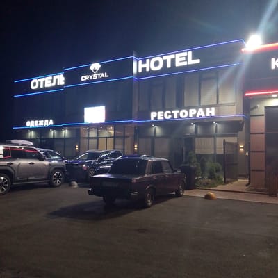 The image shows a hotel and restaurant complex at night. The building features prominent illuminated signage. On the left, the Russian word "ОТЕЛЬ" (Hotel) is displayed in white illuminated letters, followed by a logo and then "CRYSTAL". To the right, the English word "HOTEL" is displayed in large white illuminated letters. Further to the right, the Russian word "РЕСТОРАН" (Restaurant) is also in illuminated white letters. A red illuminated sign is visible on the far right.
In the foreground, several cars are parked. A silver SUV with roof rails is on the left. Next to it is a dark-colored sedan, and to the right of that is a burgundy-colored sedan with a visible license plate reading "A371УЕ 102". The ground appears to be asphalt, and there are some small plants and a fence visible behind the cars. The overall environment suggests an outdoor area adjacent to the building, likely a parking lot. The lighting is artificial, coming from the building's signs and some wall-mounted lights. There are no people visible in the image. The location is specified as Krasnooktyabrskoye, Russia.
