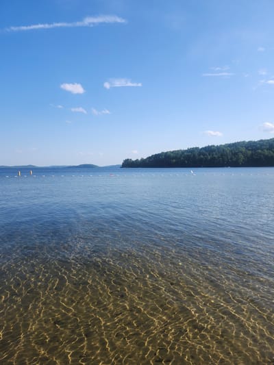 A wide-angle, eye-level shot captures a tranquil lake scene on a clear, sunny day. The foreground is dominated by the transparent, shallow water, revealing the sandy bottom with light patterns from the sun reflecting off the surface. The water gradually deepens, transitioning to a deeper blue as it extends towards the horizon. A line of white buoys with yellow markers and a red and white floating object demarcates a swimming area. In the distance, a lush, green treeline forms a dense forest that hugs the gently sloping hills of the far shore. The sky above is a vibrant blue, adorned with a few wispy white clouds. The location is identified as the public beach in Duhamel, Quebec, Canada.