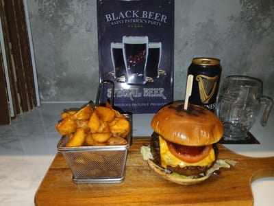 An indoor scene in Sotira, Cyprus, displays a meal and beverages on a light-colored countertop against a textured grey wall. Dominating the foreground is a light-brown wooden cutting board featuring a metal wire mesh basket filled with golden-brown potato wedges, many showing visible salt crystals. To the right of the basket, a cheeseburger is presented on the same board, secured by a wooden skewer. The burger consists of a toasted bun, lettuce, a dark patty, melted yellow cheese, a red tomato slice, and a dark red sauce. Behind the burger, a black Guinness beer can with a gold harp logo ("ESTR 1759") stands next to an empty clear glass mug with a handle, resting on a dark coaster. In the background, a rectangular metal sign depicts three glasses of dark beer, labeled "BLACK BEER" and "SAINT PATRICK'S PARTY," with "SPECIAL BEER" visible at the bottom, above partially visible text "RHEROES PROUDLY PRESENT." To the far left, a stack of brown-toned objects, possibly wooden panels or books, is partially visible. No individuals are present in the image.