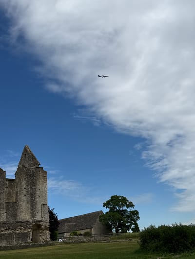 The image depicts the ruins of Minster Lovell Hall in West Oxfordshire, United Kingdom, under a partly cloudy, blue sky. A bird flies in the sky. A tree and green grass are visible to the right of the ruins.