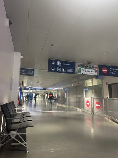 The image depicts an indoor public transit corridor within EuroAirport Basel Mulhouse Freiburg, located in Saint-Louis, France. The environment is modern, brightly lit by overhead fluorescent panels in a suspended ceiling, and features a light gray, reflective floor. A white dome-shaped security camera is mounted high on the left wall.

In the foreground on the left, a row of four black-upholstered public seating benches with metal frames is positioned parallel to the wall. A red wall-mounted fire extinguisher is visible behind these benches.

The central and right portions of the image reveal a wide corridor. Multiple overhead directional signs are present. A large blue sign displays white text and icons for "Ascenseur / Lift" with a downward arrow, and another large blue sign indicates "Information" with an 'i' symbol and downward arrows, alongside "Caisse parking / Parking Kasse / Cashier Parking" with a 'P' symbol. A banner featuring outdoor scenery and the word "Welcome" is also suspended.

On the right side of the corridor, a series of frosted or translucent modular panels with metal frames form a temporary barrier. This barrier partially conceals an area labeled "Arrivée / Ankunft" (Arrivals). Two circular red "no entry" signs, translated as "Passage interdit / Kein Zutritt," are attached to the lower section of these barriers.

Several individuals are present in the corridor. One person, wearing dark clothing and carrying a reddish-orange bag, is walking towards the camera from the mid-ground on the right side of the corridor. Further in the background, a small group of indistinct figures is visible, with one individual appearing to be seated on a bench or chair. No direct interactions between individuals are discernible.