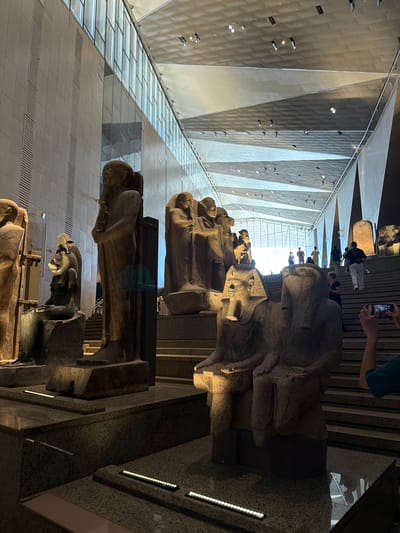 The image depicts an interior view of a large, modern museum or architectural space. The primary subjects are several large, ancient-looking stone sculptures, possibly statues or sarcophagi, arranged vertically in the frame. These sculptures are detailed, with intricate carvings suggesting human or animal forms, and they appear to be made of stone with a weathered texture.

In the background, a grand staircase with polished stone steps is visible, leading upwards. Several people are present on the staircase, some looking at the sculptures, others taking photographs with their phones, indicating a public visiting the exhibit. The architecture features a dramatic, angular ceiling with metallic or reflective panels and integrated lighting, creating a sense of scale and grandeur. The lighting suggests it's daytime, with natural light entering from a high point in the ceiling, illuminating the space and the sculptures.

The scene conveys a sense of awe and appreciation for the historical artifacts within a contemporary architectural setting. The sculptures are the clear focal point, evoking a connection to ancient history. The presence of visitors engaged with their phones and looking at the exhibits suggests a lively and accessible cultural experience. There is no visible text in the image. The location is contextually identified as Kafrat Nassar, Egypt.