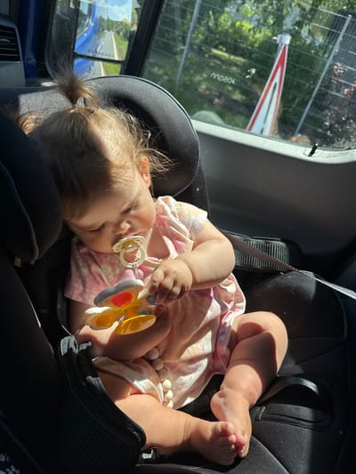 A young child is seated in a car seat, facing forward. The child has blonde hair pulled into a small ponytail and is wearing a pink dress with a floral pattern. The child is holding a yellow and red toy and has a pacifier in their mouth. The car seat is black and secured with a seatbelt. Sunlight streams in from the right side of the image, casting shadows. Through the window behind the child, a road is visible, with trees and a tall, striped marker on the right. The interior of the vehicle appears to be a van or bus, with a vent visible on the left side. The exterior scene suggests a daytime setting, possibly in a suburban or rural area. The image was taken in Oberwetz, Germany.