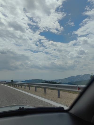 The image captures a view from a vehicle driving on a road, likely a highway, on a cloudy day. In the foreground, the dashboard of the vehicle is visible, obscuring the lower portion of the scene. The road itself is paved with asphalt and has a white lane marking. To the right of the road, a metal guardrail with reflective markers separates the roadway from the embankment. The embankment appears to be covered in sparse vegetation and dirt.
In the middle ground, rolling hills and mountains stretch across the horizon, some covered in greenery, others appearing rocky. The sky is filled with a mix of white and grey clouds, with patches of blue sky visible. The overall atmosphere suggests a journey through a natural, possibly mountainous, landscape. No people or specific landmarks are identifiable that would pinpoint the location within Komotini, Greece, but the scene depicts a typical road environment outside of a city center, likely in a rural or semi-rural area.