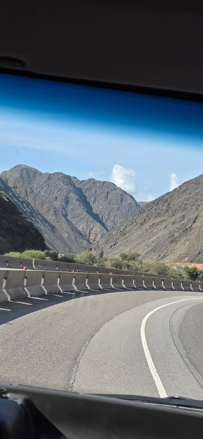 The image is taken from inside a vehicle, looking out at a mountain landscape. A curved asphalt road with a white line marking the lane is visible in the foreground, bordered by a concrete barrier with red markers. Beyond the barrier, sparse green vegetation grows on the mountainside. In the background, rugged, arid mountains rise under a bright blue sky with scattered white clouds. The scene suggests a journey through a mountainous region, possibly a highway passing through a valley. There are no people or specific landmarks identifiable that would pinpoint a location within the city of Sasyk-Bulak, Kyrgyzstan.