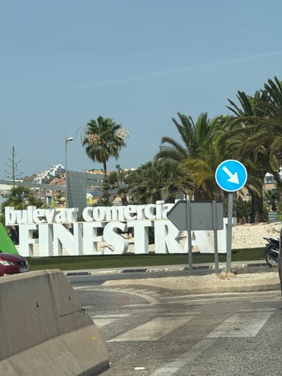 This is a photograph taken outdoors on a clear, sunny day. The foreground features a concrete barrier with a textured surface and some visible damage. Beyond the barrier, a street with a pedestrian crossing is visible, showing signs of wear and tear. A red car is parked to the left, with a license plate that reads "3910 HNC".

In the mid-ground, a large, white, three-dimensional sign displays "buLeVar COMERCIa" on the top line and "FINESTR" on the bottom line. The letters are bold and blocky, set against a backdrop of green Astroturf. Behind the sign, palm trees of various sizes are prominent, with lush green fronds. A tall, slender street lamp is visible to the left of the sign.

Further in the background, a hillside is dotted with white buildings with red-tiled roofs, suggesting a coastal or Mediterranean setting. A blue circular sign with a white arrow pointing to the right is mounted on a pole. Another grey directional sign with an arrow pointing to the left is also present. In the far right of the image, a portion of a motorcycle is visible. The overall environment suggests a location in a warm, sunny climate. The sign indicates the location is likely a commercial boulevard in a place named "Finestrat". Given the visual cues of palm trees, white buildings, and clear blue sky, it is plausible this is La Cala, Spain, a coastal town known for its beaches and tourist amenities.