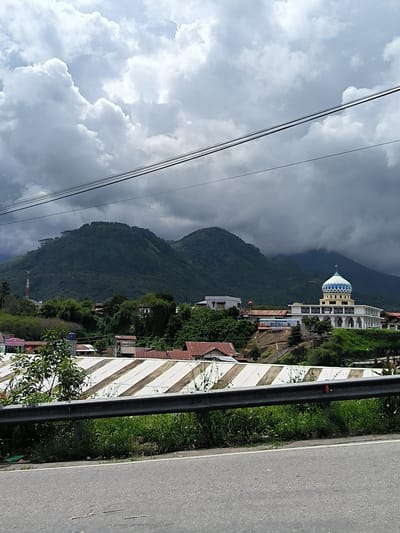 In the city of Babussalam, Indonesia, a mosque with a blue and white domed roof stands prominently on a hill. The mosque features white walls with arched windows and a distinctive minaret. Surrounding the mosque are several buildings, including some with red tiled roofs, and lush green vegetation. In the foreground, a grey metal guardrail runs along the side of an asphalt road. The road is bordered by green grass and some litter. The background is dominated by a large, green, tree-covered mountain range, partially obscured by dramatic, dark grey clouds. Power lines are visible strung across the sky.