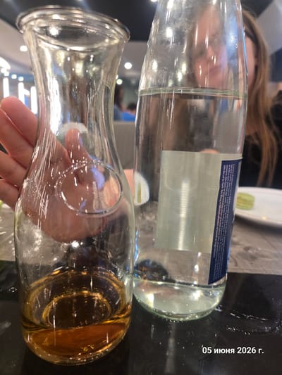 On June 5, 2026, in La Louvière, Belgium, a close-up indoor scene depicts two glass containers on a dark, reflective surface. The container on the left is a clear glass carafe, approximately one-fifth full of an amber-colored liquid. A person's hand, featuring light skin tone and a pink-painted fingernail, grasps the carafe at a mid-section indentation. The container on the right is a clear glass bottle, approximately one-third full of clear liquid, with a vertical label partially visible, containing text in French and German. Small water spills are present on the dark surface to the right of the bottle. In the blurred background, a person with long, light brown hair and dark clothing is partially visible through the glass bottle. Additional highly blurred figures and illuminated circular light sources are visible in the far background, suggesting a public dining establishment. A white plate with a small green object (possibly a macaron) is indistinctly visible in the far right background.