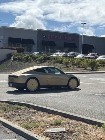 The image depicts a gold-colored concept car parked in a parking lot outside a Planet Fitness gym in San Bruno, California, USA. The car has a futuristic design with a sleek, aerodynamic body and solid wheel covers. In the background, the Planet Fitness building is visible with its distinctive purple logo and signage. Several other cars are parked in the lot behind the concept car. The scene is set on a sunny day with scattered clouds in the sky.