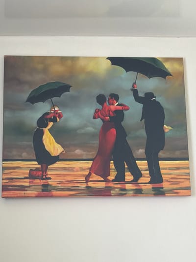 The image is a painting of four figures in a moody, overcast outdoor setting, likely a beach or waterfront due to the wet, reflective ground and the presence of umbrellas.
On the left, a figure dressed in a black dress and yellow apron, holding a green umbrella, appears to be struggling against the wind, with their body bent and their head tilted. A red bag rests on the ground beside them.
In the center, a couple is embracing and dancing. The woman wears a striking red dress, and the man is in a dark suit. They are positioned under the open sky, with no umbrella.
On the far right, a man in a dark suit and hat holds a dark green umbrella over himself and a partially visible figure. He is holding a pale object, possibly a hat or a scarf, in his other hand.
The ground is rendered with a high degree of reflectivity, suggesting it is wet from rain or sea spray, with distorted reflections of the figures and umbrellas visible. The background depicts a dramatic sky with swirling clouds in shades of grey, yellow, and hints of blue, adding to the somber and atmospheric mood of the scene.
The painting's style is illustrative and somewhat stylized, with a focus on capturing movement and emotion. The location in Uhersko, Ukraine is not visually evident in the painting itself.