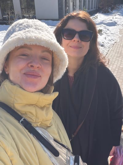 The image displays two adult females outdoors on a clear, sunny day in Ракаўскі сельскі Савет, Belarus. The woman in the foreground, left, has light brown hair, wears a white fluffy bucket hat, a yellow puffer jacket, and a silver-colored shoulder bag with a black strap. She is looking at the camera with a slight smile. The woman on the right, positioned slightly behind, has dark brown hair, wears dark sunglasses, and a dark coat over a reddish-brown knitted top. She is also looking towards the camera with a slight smile. The background features modern buildings with large windows, patches of snow on the ground next to a paved walkway, and bare trees. The scene is brightly illuminated by direct sunlight.
