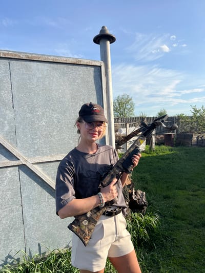 A person wearing glasses and a black baseball cap is holding a crossbow. The person is wearing a gray t-shirt and light-colored shorts. They are standing next to a metal structure with a post extending upwards. Behind the person is a grassy area and a wooden structure. The sky is blue with some clouds. The image was taken in an outdoor setting. Given the prompt, the location is assumed to be in or near Слобода, Russia.
