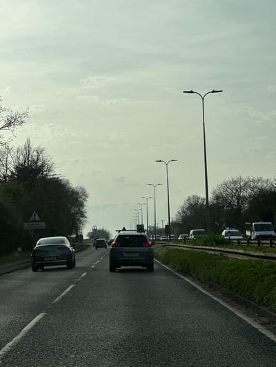 The image depicts an asphalt road scene in Basildon, United Kingdom, specifically on Laindon Link (B1007) heading northbound towards the Fortune of War roundabout and the A127 (Southend Arterial Road). The sky is overcast with visible cloud formations.
In the foreground, a dark grey Peugeot 3008 SUV, bearing the registration SR70 ZPE, is visible with roof racks, facing away from the viewer. Immediately behind it, a dark grey sedan with registration BG70 XOW is also traveling in the same direction. Multiple additional vehicles, including cars and vans, are present in the mid-ground and background, indicating moderate traffic flow.
On the left side of the road, a triangular warning sign with a red border and a black pedestrian symbol is visible, below which a rectangular white sign reads "Pedestrians Crossing Ahead". The left roadside features dense vegetation, primarily deciduous trees with bare branches or nascent foliage. A prominent yellow McDonald's 'M' arch logo is visible in the mid-distance, positioned to the right of the road's center line.
The right side of the road features a grass verge, a low metal barrier, and further bare-branched trees and sparse vegetation. A series of modern streetlights, each with a dual fixture, extends along both sides of the road into the distant background, indicating a major arterial route. No pedestrians are visible in the scene.