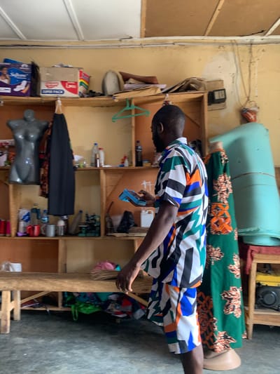 The image depicts an indoor scene, possibly a workshop or shop, in Jos, Nigeria. A man wearing a colorful, geometric-patterned shirt and shorts stands in the foreground, holding a small blue object in his right hand. He is facing away from the camera, towards a shelf unit. The shelves are wooden and appear to be stocked with various items, including bottles, containers, fabric, and a sewing machine. To the left of the man, a dark grey mannequin torso is visible. To his right, a mannequin is dressed in a green, floral-patterned dress. Behind the mannequins, a large, light blue cylindrical object is present. The walls of the room are painted a light yellow, and the ceiling is white with visible beams. The floor is a dark grey concrete. The overall impression is that of a busy, functional space, likely involved in textile work or retail.