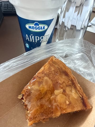 The image depicts a close-up, elevated view of a snack arrangement on a dark brown surface, consistent with a desk, in Senokos, Bulgaria. A triangular slice of pastry, approximately 15 cm along its visible longest edge, occupies the foreground. The pastry features a golden-brown, glossy top layer embedded with numerous almond slivers. A bite has been taken from one corner of the pastry, exposing a reddish-brown filling beneath the crust. The pastry rests on a piece of brown cardboard packaging, partially covered by a transparent plastic bag. Behind the pastry, a tall, white and dark blue plastic cup is visible, prominently displaying the "MEGGLE" logo in green and the Cyrillic word "АЙРЯН" (Ayran) in white. To the right of the cup, a clear glass with a distinct vertical fluted pattern is partially visible. In the upper left background, an out-of-focus black object with a grid-like texture, resembling a computer keyboard or similar electronic device, is discernible. No individuals are present in the scene.