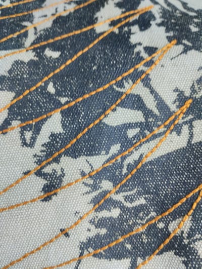 The image is a close-up shot of a textured fabric with a dark gray and white pattern, overlaid with thin, parallel orange stitches. The fabric appears to be a canvas or a similar coarse material, with visible woven threads creating a subtle grid. The pattern on the fabric is abstract, resembling splashes or organic shapes in dark gray against a lighter background. The orange stitches run diagonally across the frame from the bottom left to the top right, creating a series of equidistant lines. The lighting is even, and the focus is sharp on the fabric's texture and the stitching. There are no people, animals, or identifiable landmarks visible in the image. The image does not provide any information that would allow for its location to be specified within Castione Andevenno, Italy, or any other geographical area.