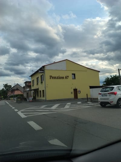 The image shows a street scene in Opařany, Czechia, under a cloudy sky. On the right side of the road, a bright yellow building with the text "Penzion 67" is visible. A white car is parked in front of this building. The street has white lane markings and a pedestrian crossing. In the background, there are other buildings and trees. The overall impression is of a quiet, possibly rural, setting.