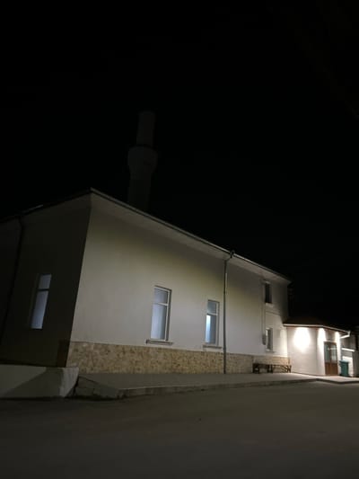 The image depicts a mosque at night in Hvostiane, Bulgaria. The central building is a single-story structure with white-painted walls, featuring a lower facade section clad in light-toned, irregular stone masonry. A slender, cylindrical minaret with a narrow cap rises from the roofline on the left. The main building's facade contains three rectangular windows, illuminated from the interior, casting soft light outwards. A silver-colored downspout runs vertically along the right side of the main building, below a matching roof gutter. To the right, an attached section features a brown, overhanging roof supported by rafters, an exterior wall-mounted light fixture, a door, and an additional window. A plain wooden bench is positioned on the paved sidewalk in front of this attached section, alongside a dark green refuse bin. The asphalt street in the foreground is unlit and vacant. No individuals or vehicles are present in the scene.