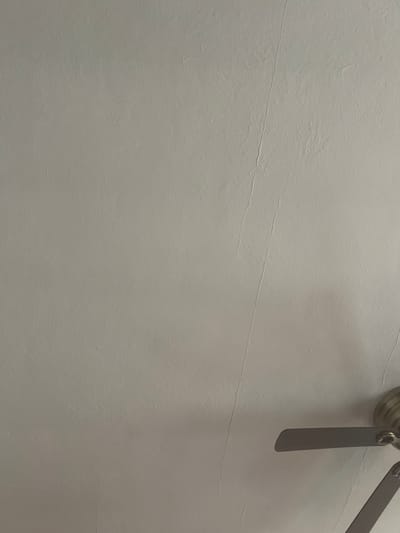 The image shows a close-up view of a textured white ceiling with a faint crack running vertically down the center. A portion of a ceiling fan, with brass-colored housing and dark blades, is visible in the lower right corner. The scene appears to be an indoor residential setting, likely a bedroom or living room. The lighting is dim, casting a shadow from the fan onto the ceiling. No people are present in the image. The location is identified as West Hartford, United States of America.