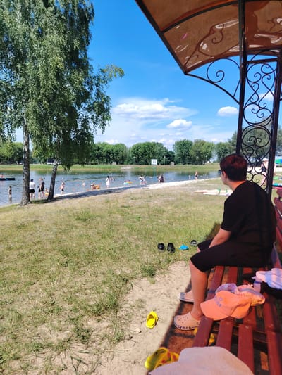 The image depicts a crowded beach scene on a sunny day, likely in Malaryta, Belarus. In the foreground, a young person wearing a black t-shirt and black shorts sits on a wooden bench, facing away from the camera and looking towards a lake or river. Their feet are resting on the ground, and they are wearing white Crocs. Several pairs of shoes and sandals are scattered on the ground and on the bench, along with some clothing items.
The middle ground of the image shows the shore of a body of water, with a sandy and grassy area. People are swimming, wading, and relaxing in and around the water. Some are playing with inflatable rings. A few trees stand on the grassy bank. In the background, a line of trees forms the horizon, and behind them are buildings, suggesting a proximity to a town or settlement. The sky is bright blue with scattered white clouds. The overall atmosphere is one of recreation and summer leisure.