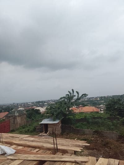 This image captures an elevated view of a residential area in the city of Arun, Nigeria, under a uniformly overcast, grey sky. In the foreground, a large, stacked pile of light-colored wooden planks and beams occupies the lower portion of the frame, indicating construction materials. Vertical metal rebar rods are visible protruding from within or near the lumber, and a white sack, possibly containing building aggregates, is situated on the left. Directly behind these materials, a small, rudimentary single-story wooden shack with a corrugated metal roof stands, partially enveloped by green foliage from a tree. A light-colored concrete block wall with a red metal gate is visible to the left of the shack. The midground and background present a sprawling urban landscape of closely packed residential buildings with diverse roof types, predominantly terracotta/orange and some green/grey, extending into the distant horizon across gently rolling terrain. No individuals are present, and no overt actions or interactions are depicted within the scene.