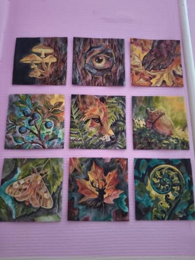 The image displays a collection of nine small, square watercolor paintings arranged in a 3x3 grid on a pink, textured surface. The paintings depict various elements of nature. The top row features mushrooms, a stylized eye within a tree trunk, and a bear's paw among autumn leaves. The middle row shows blueberries on a branch, a fox peeking through ferns, and an acorn. The bottom row includes a moth, a deer silhouette on a maple leaf, and a fern frond. The location of this image is not specified as being in Kyiv, Russia, as that is a geographic impossibility.
