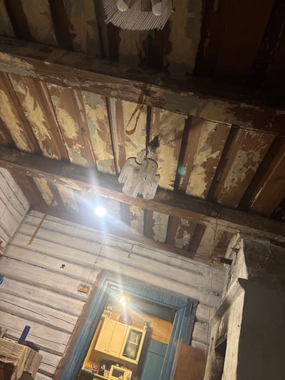 The image captures an interior scene of what appears to be an old, rustic dwelling. The viewpoint is looking upwards towards the ceiling and a doorway. The ceiling is constructed of wooden planks with visible beams, and the paint is peeling, revealing older layers of color and bare wood. Two chandeliers are visible: one at the top of the frame, a modern fixture with a textured cover, and another more ornate, crystal-like chandelier hanging lower down, illuminated by a bright light source. The walls are made of horizontally stacked logs, painted white with visible cracks and imperfections.

In the lower part of the image, a doorway with a blue frame leads into another room where kitchen cabinets and a microwave are visible. The lighting is dim and atmospheric, with a bright light source emanating from the lower chandelier and a smaller, circular light fixture on the wall. There are electrical outlets on the log wall near the doorway. A piece of fabric, possibly a tablecloth, is draped over a surface in the foreground on the left. A thin wire hangs vertically from the ceiling towards the center of the frame. The overall impression is one of a weathered, perhaps abandoned or simply old, rural home. The location in the Shatsky rural council, Belarus, suggests a setting consistent with traditional Belarusian rural architecture. No people or animals are visible in the image.