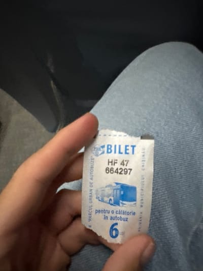 The image is a close-up shot captured in Cricova, Moldova. A fair-skinned left hand is shown holding a white, slightly crumpled paper bus ticket. The ticket displays blue text and a graphic of a city bus. Printed details include the word "BILET" (ticket), the ticket number "HF 47 664297," the issuing authority "PRIMĂRIA MUNICIPIULUI CHIȘINĂU" (Chisinau Municipality City Hall) and "PMA PARCUL URBAN DE AUTOBUZE" (Urban Bus Park). It states "pentru o călătorie în autobuz" (for a bus ride) and indicates a fare of "6 LEI" (Moldovan Leu). The hand and ticket are positioned over a lap covered by light blue denim jeans, visible in the lower right portion of the frame. The upper background is a dark, blurred area, consistent with the interior of a vehicle's seating environment.
