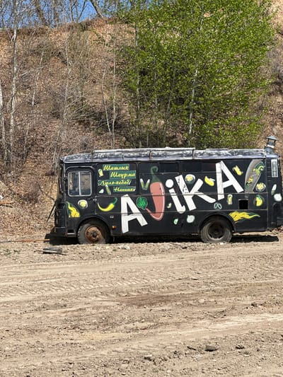The image depicts a black food truck parked on a dirt road in what appears to be a wooded area. The truck has a large, white inscription on its side, along with images of peppers, herbs, and flames. There are also Cyrillic words, which translate to "Shashlik, Shawarma, Lyulya-kebab, Drinks". The location is identified by a sign on the truck as Chigiri, Russia.
