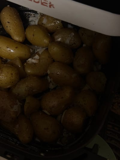 The image shows a close-up view of roasted potatoes in a black cooking pan. The potatoes are small and oval-shaped, with golden-brown skin and visible seasoning. Some pieces of garlic are interspersed among the potatoes. The lighting is dim, with the upper part of the pan slightly obscured by a white appliance. The background is dark and out of focus, with some hints of a wooden surface. There are no people or distinct urban elements visible in the image, making it impossible to determine a specific location in Cluses, France.
