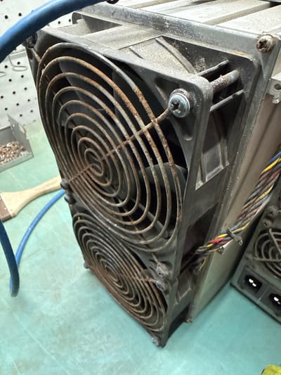 The image shows a close-up, low-angle view of two large, dusty computer cooling fans mounted on industrial equipment, possibly part of a cryptocurrency mining rig. The fans have rusty grilles and are secured with visible screws. A bundle of colored wires is tied with black zip ties, emerging from the side of the equipment. A blue electrical cable snakes across the foreground, and a paintbrush with a wooden handle lies on a light green, textured surface in the background. The overall scene suggests a workshop or industrial setting. No specific location in Indiantown, United States of America, is discernible from the image itself.