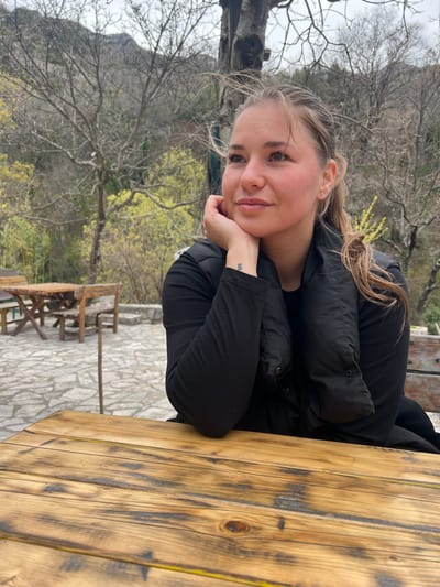 A woman is resting her head on her hands while sitting at a wooden table outside. She is wearing a black shirt and a black vest. There are trees and mountains in the background. The location appears to be a restaurant in a rural area, potentially in the city of Krapina, Montenegro. A wooden picnic table and chairs are visible in the background. The ground is paved with stone tiles.