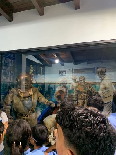 In Boca del Río, Venezuela, a group of people, primarily children with dark hair, are looking at a museum exhibit through a glass display case. The exhibit features mannequins dressed in vintage diving suits. One mannequin in the foreground is wearing a copper helmet and a tan suit with a gray pouch. Behind it, other mannequins depict divers in different stages of their gear, some with hats. The display includes historical photographs and text panels on the back wall, one of which appears to be a painted mural of divers and historical equipment, including a scale. The environment is dimly lit, with wooden beams visible on the ceiling. The overall scene captures a moment of learning and observation at a historical exhibit.