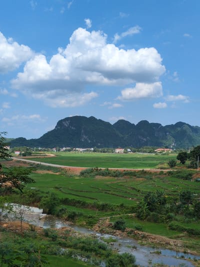 The image captures a scenic view of a rural landscape in Ngọc Lặc Commune, Vietnam. The foreground features a meandering river with rocky banks, surrounded by lush green vegetation and trees. A dirt road curves through the middle ground, separating a terraced rice field from a broader expanse of green fields. In the background, a cluster of houses forms a small village nestled at the base of a dramatic, verdant mountain range that dominates the horizon. The sky above is a vibrant blue, dotted with fluffy white clouds, suggesting fair weather. The overall impression is one of tranquility and natural beauty.