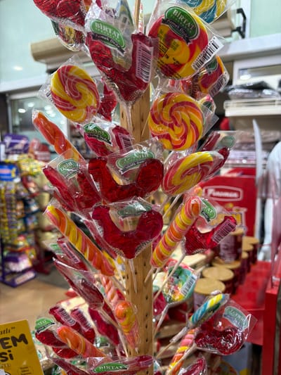 This image shows a close-up view of a display rack filled with a variety of colorful lollipops and candies. The candies are individually wrapped in clear plastic packaging, with some displaying branding that includes the text "BİRTAT Pamuk Dede". The rack appears to be made of light-colored wood. In the background, shelves are visible stocked with various products, suggesting this is a retail environment. The scene is likely located in the city of Acıpayam, Turkey, given the specific branding and general context of a commercial establishment. There are no people visible in the immediate frame.