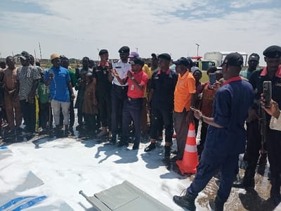 The scene is a public space in Katabu, Nigeria, located at coordinates 10.71 latitude and 7.514 longitude. There is a large crowd of people gathered around a white foamy substance on the ground. The substance appears to be some kind of foam or detergent. There are several men in uniform, some wearing black and some wearing red and white, standing among the crowd. They are interacting with the group, talking and gesturing. One man in blue overalls stands with his back to the camera, facing the crowd, and is holding a smartphone in his right hand. The scene is a public event, possibly a demonstration or a product launch. There are no visible signs or other identifying features that help to determine the event's purpose.