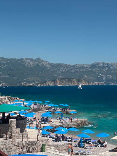 The image depicts a vibrant coastal scene in the city of Kubasi, Montenegro, under a clear, bright blue sky. In the background, a range of rugged, forested mountains with visible rock formations stretches across the horizon, with scattered settlements discernible on their lower slopes. The deep blue sea occupies the middle ground, transitioning to a clearer turquoise closer to the shore. A white sailboat with its sails unfurled is visible on the open water.
The foreground features a multi-tiered beach area constructed with natural stone walls and pathways. Numerous light blue beach umbrellas provide shade over rows of white lounge chairs with blue cushions, occupied by individuals. People are seen relaxing, sunbathing, and walking on the beach terraces. On the left, a prominent stone structure with a green canopy, likely a beach bar or restaurant, hosts several patrons. Further right and closer to the water, multiple individuals are swimming or wading in the clear, shallow sea. A green trash bin is visible on one of the lower stone terraces.